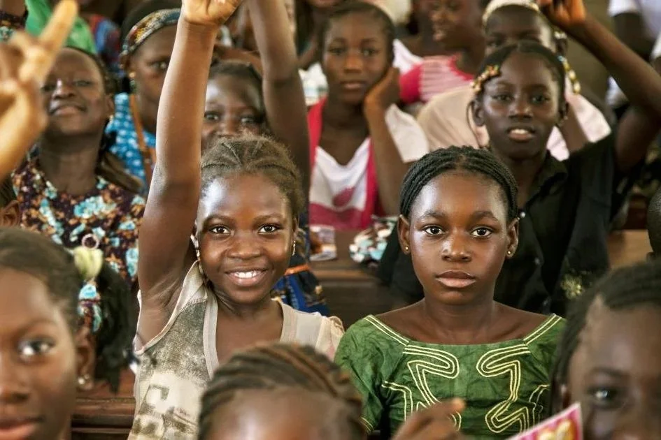 A group of young girls sitting in a classroom, with one girl raising her hand, looking engaged and attentive.