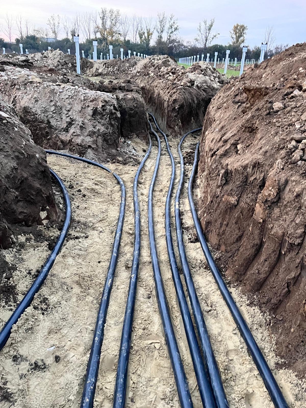 Multiple black electrical cables laid in a trench during construction, with dirt and soil on either side, and trees in the background.