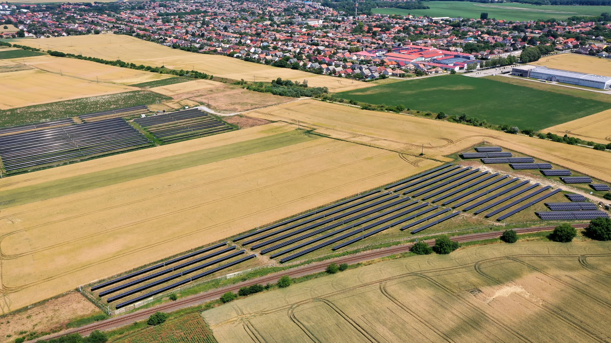 Aerial view of a solar farm with multiple rows of solar panels situated in agricultural fields near a town with residential neighborhoods and some industrial buildings.