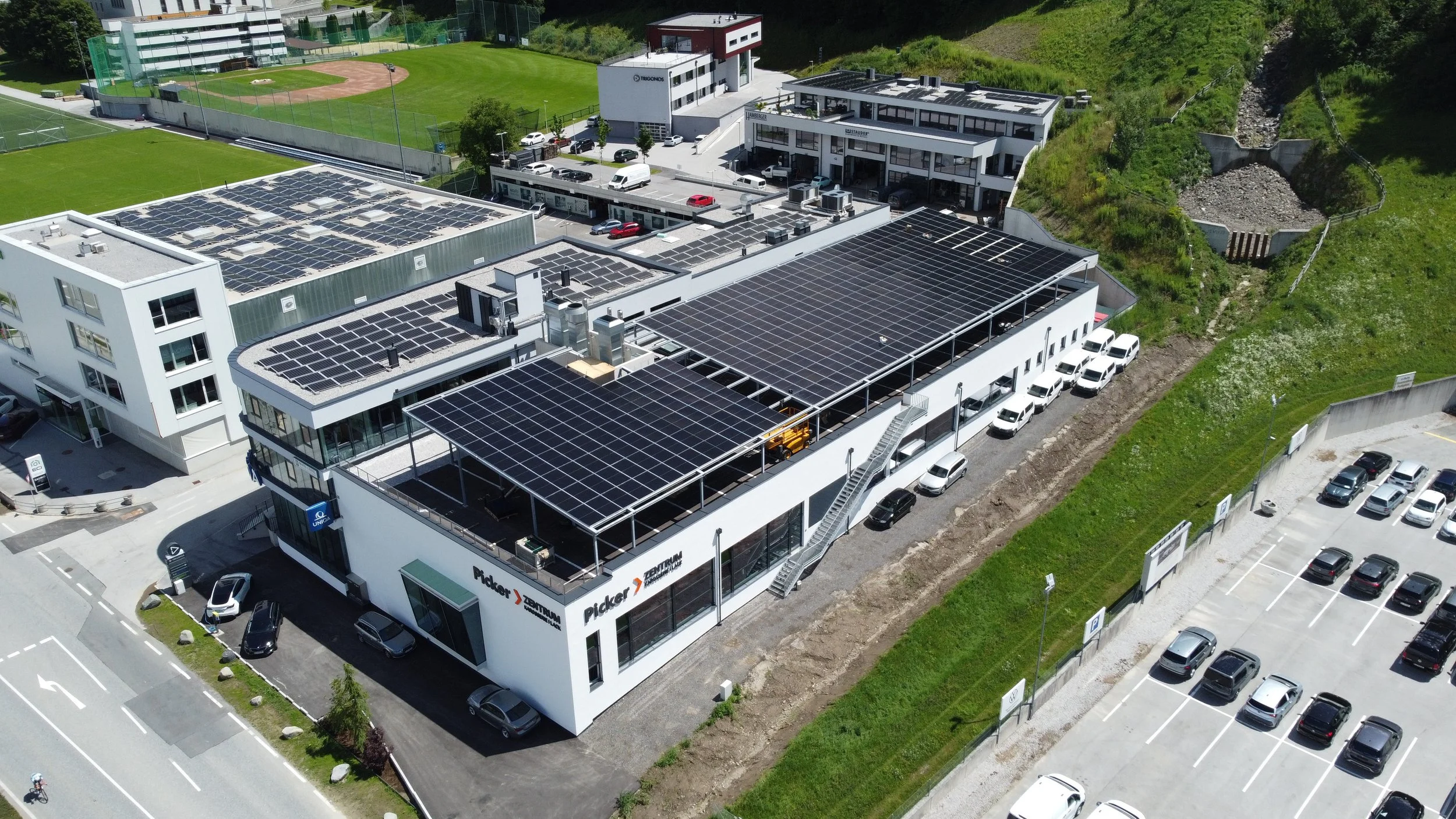 Office building with solar panels on roof, parking lot, adjacent sports field, and green hillside with erosion control structure.