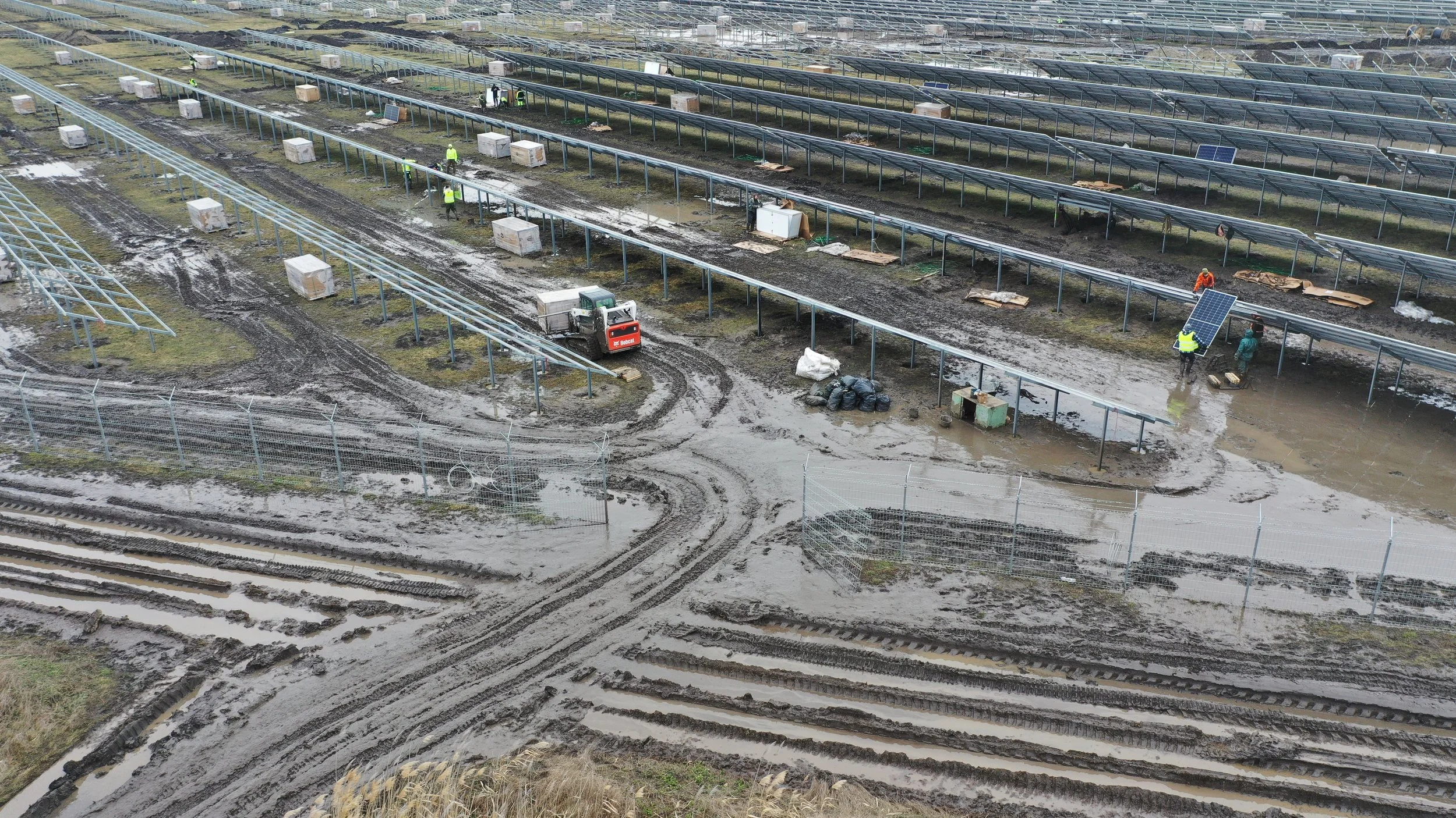 Workers installing solar panels on muddy ground in a solar farm, with some panels already installed and others being prepared for installation.