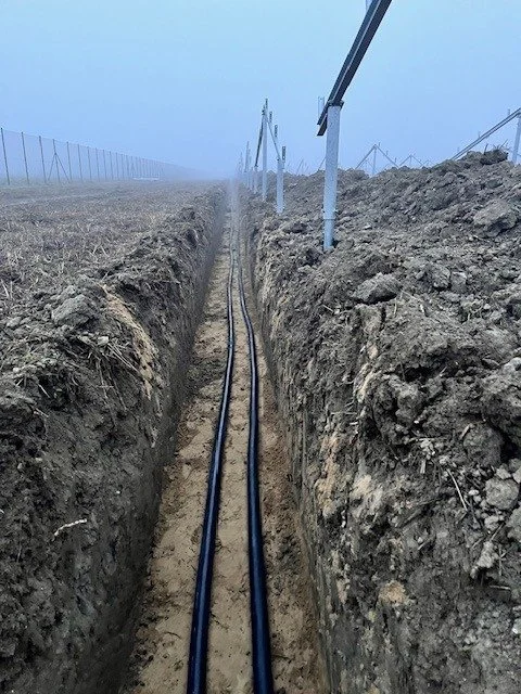 Trench with two black cables installed in the ground, supported by metal brackets on a foggy day in an open field with fencing.