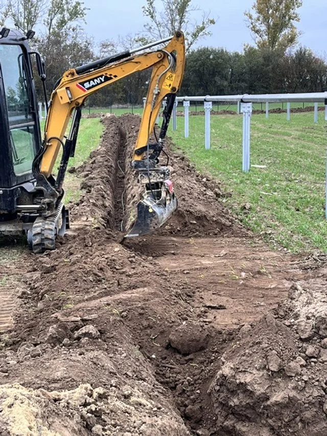 A yellow and black SANY mini excavator digging a trench in the ground next to a grassy field and a white fence.