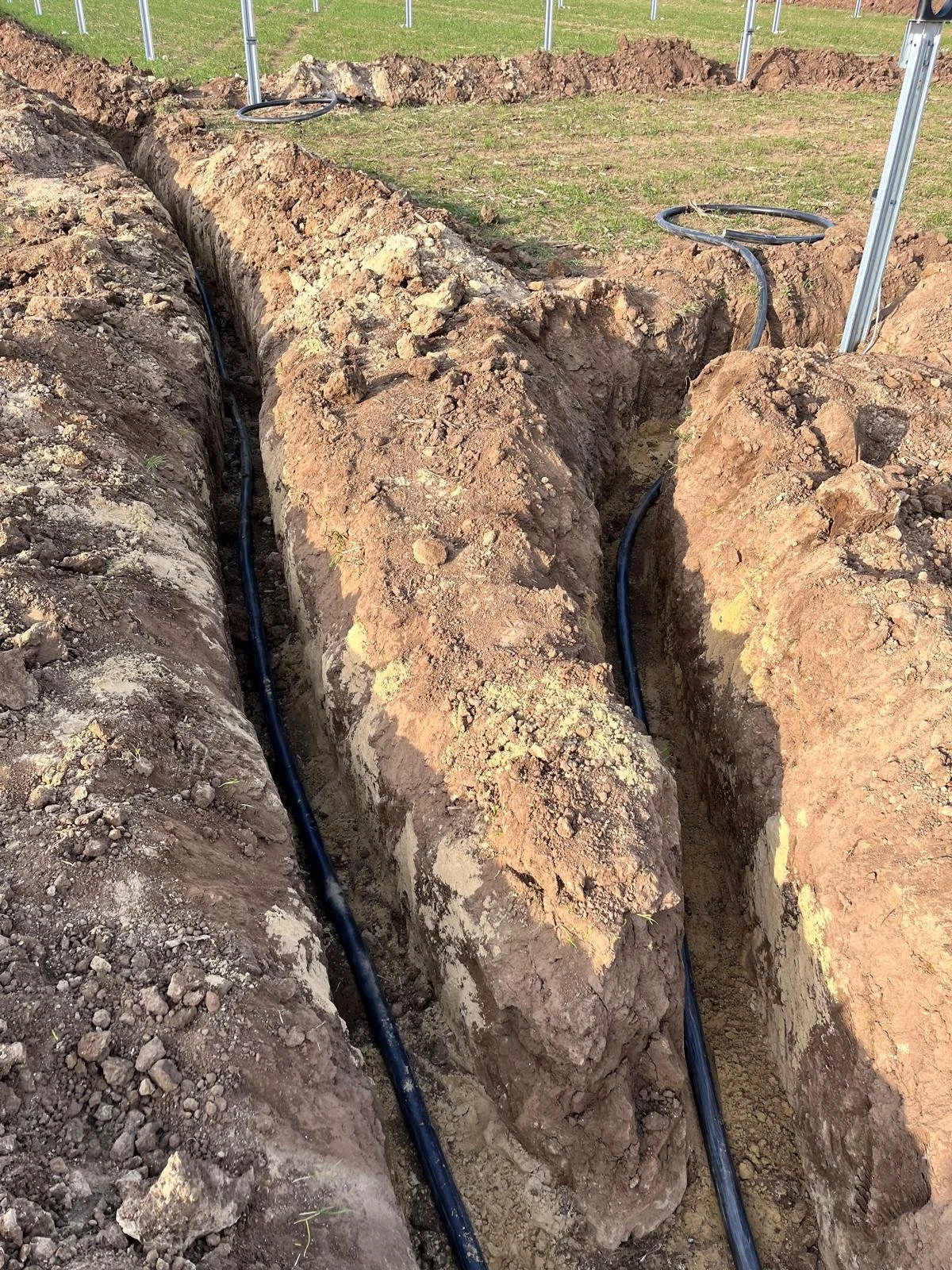 Trenched soil with black underground cables being installed in a field with metal poles, likely for electrical or communication utilities.