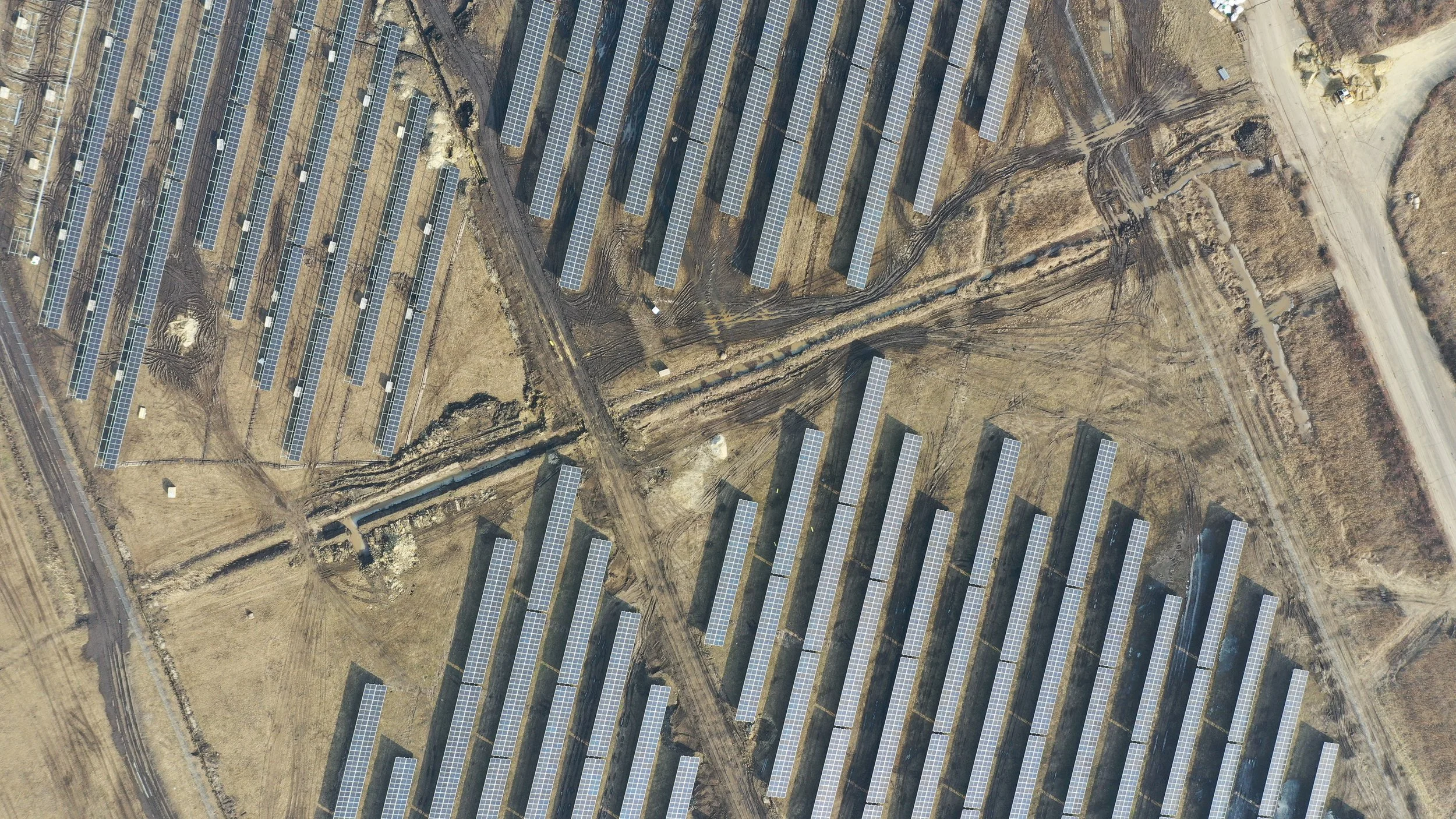 Aerial view of a solar farm with multiple rows of solar panels on a dry, grassy landscape, intersected by dirt pathways and adjacent to a paved road.
