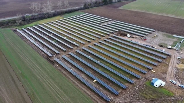 An aerial view of a solar farm with multiple rows of solar panels on a large field, some construction activity, and a small white building nearby.