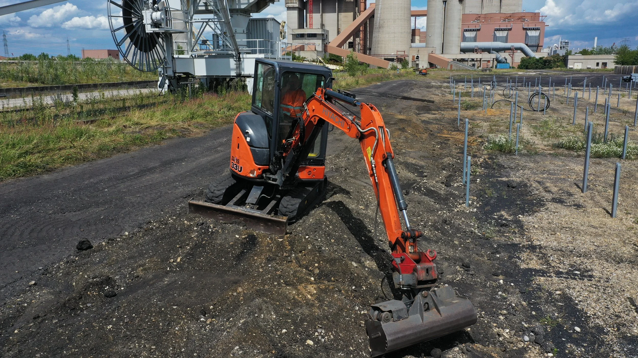 Egy kis narancssárga-fekete minikotrógép dolgozik egy építési területen, ipari üzem háttérrel