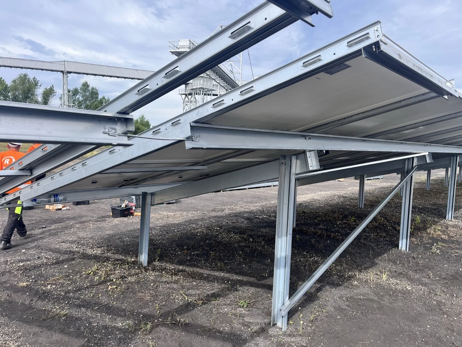 Metal platform structure with support beams and stairs, outdoors on dirt ground, with a person in safety vest and helmet nearby, and industrial equipment and sky in background.