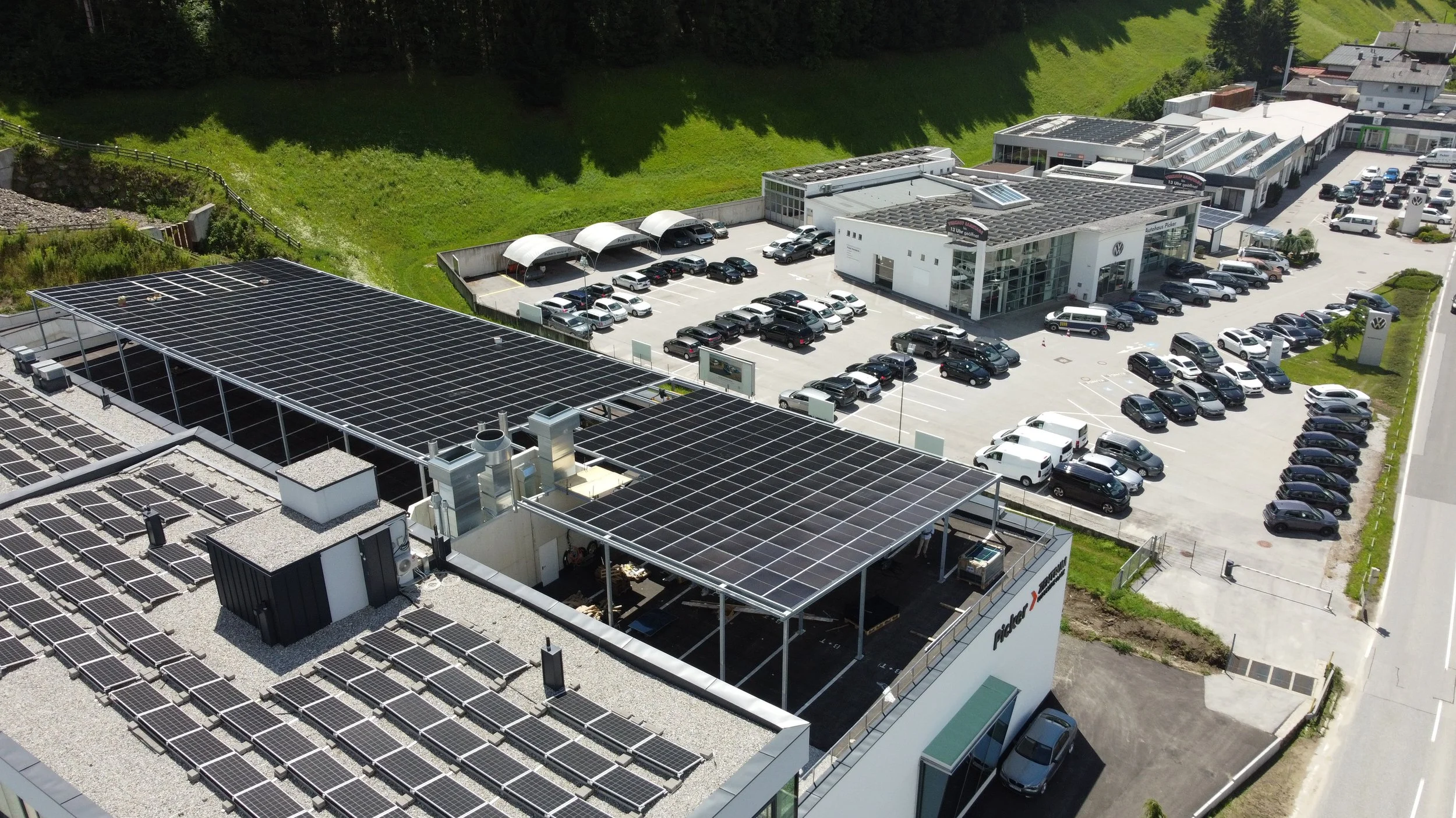 A building with solar panels on the roof, adjacent to a parking lot filled with various cars in front of a Volkswagen dealership, surrounded by green landscape and other commercial buildings.