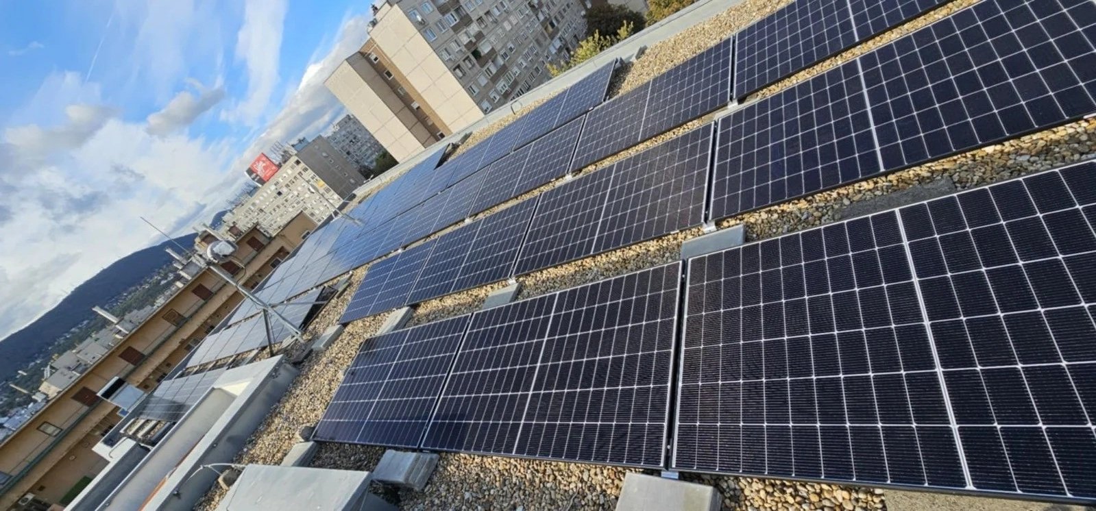 Multiple solar panels installed on a building rooftop against a backdrop of city buildings and mountains.