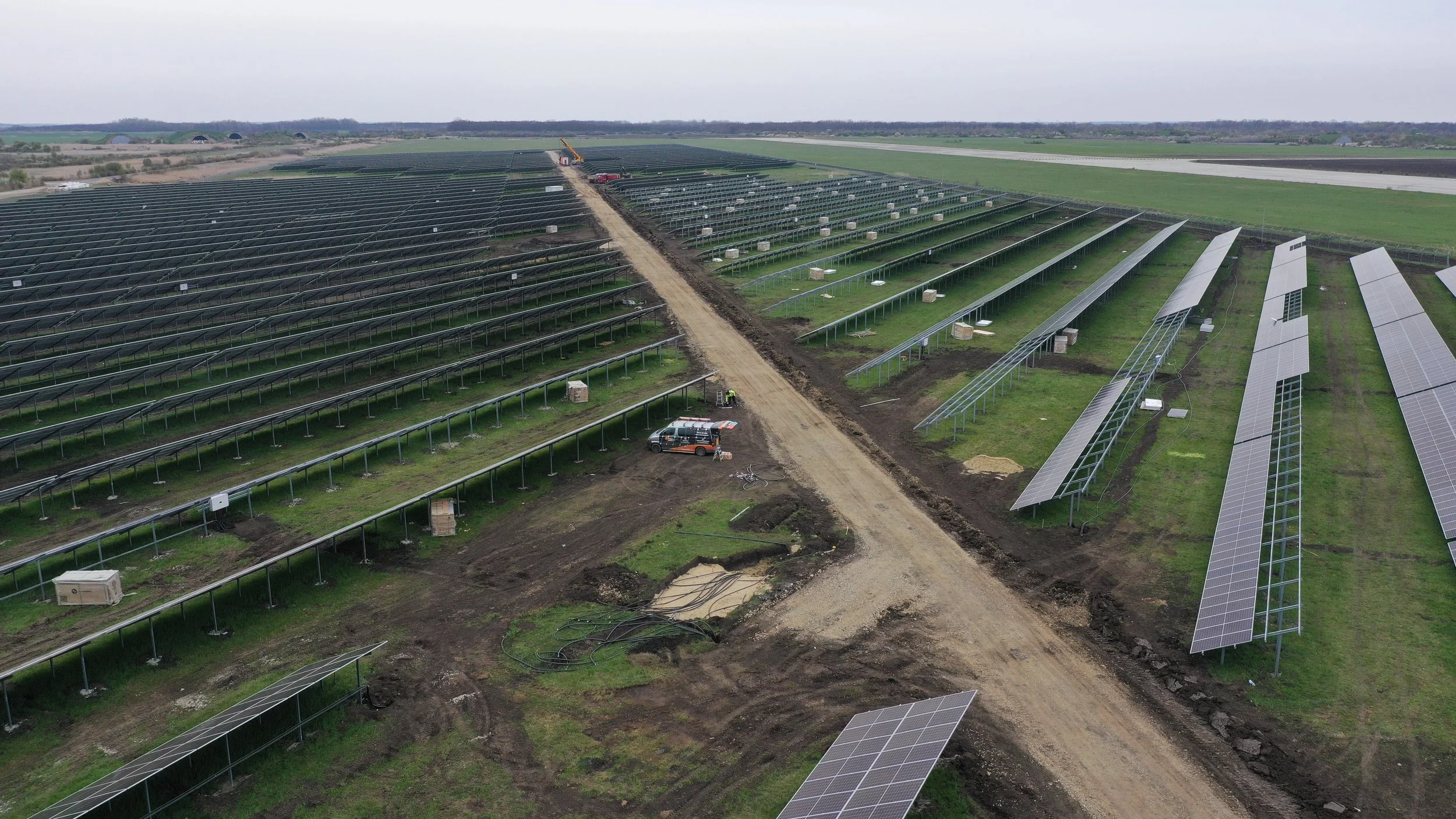 A large solar farm under construction with numerous solar panels being installed in an open field, a dirt road running through the site, construction vehicles and equipment visible.