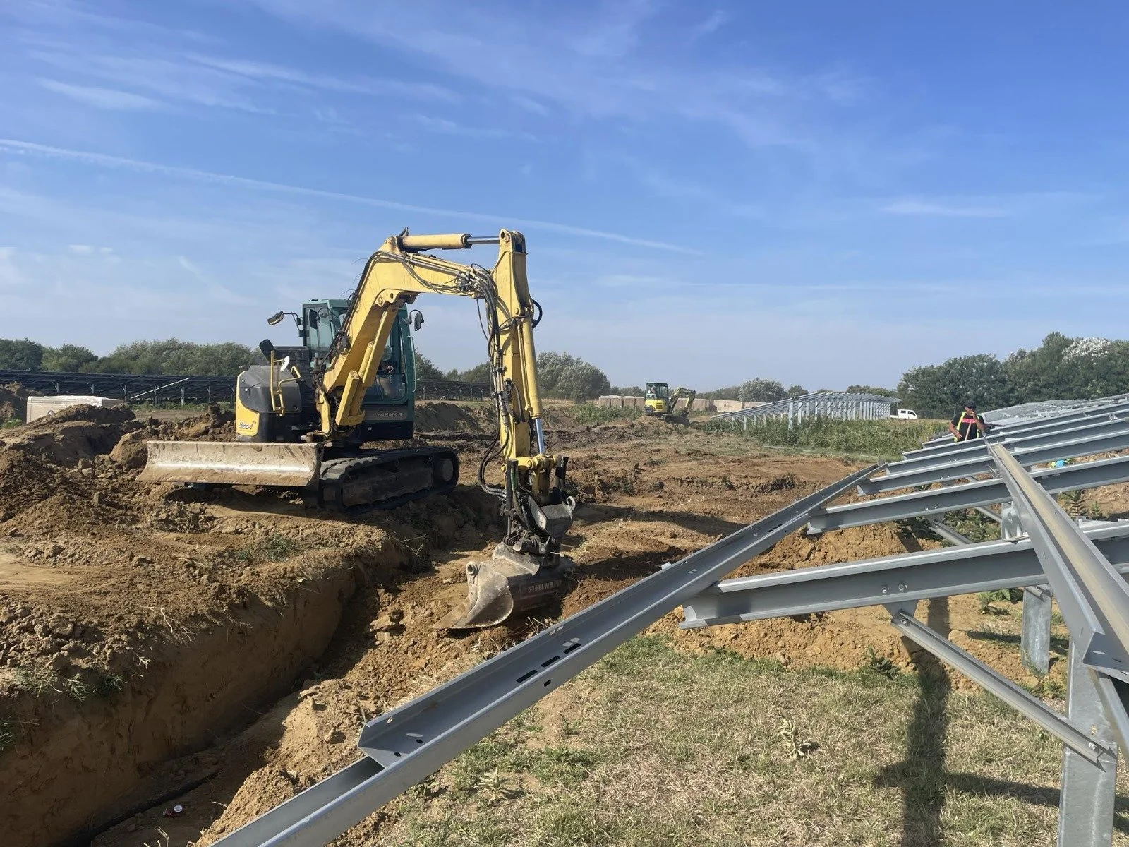 Construction site with excavator digging a trench and metal framework structure nearby on a sunny day.