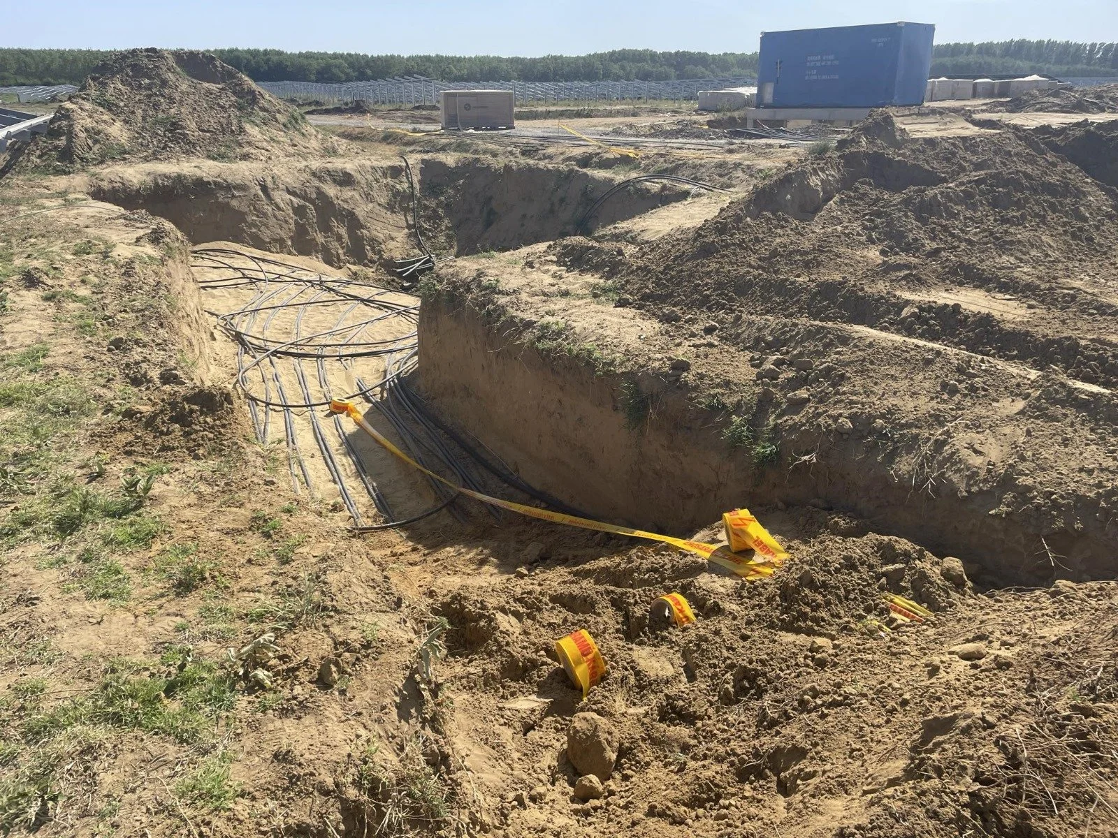 Construction site with underground electrical or communication cables laid in a trench, surrounded by mounds of dirt and yellow caution tape.