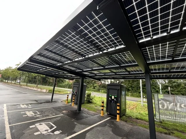 Electric vehicle charging station with solar panels overhead, situated in an outdoor parking lot surrounded by trees.