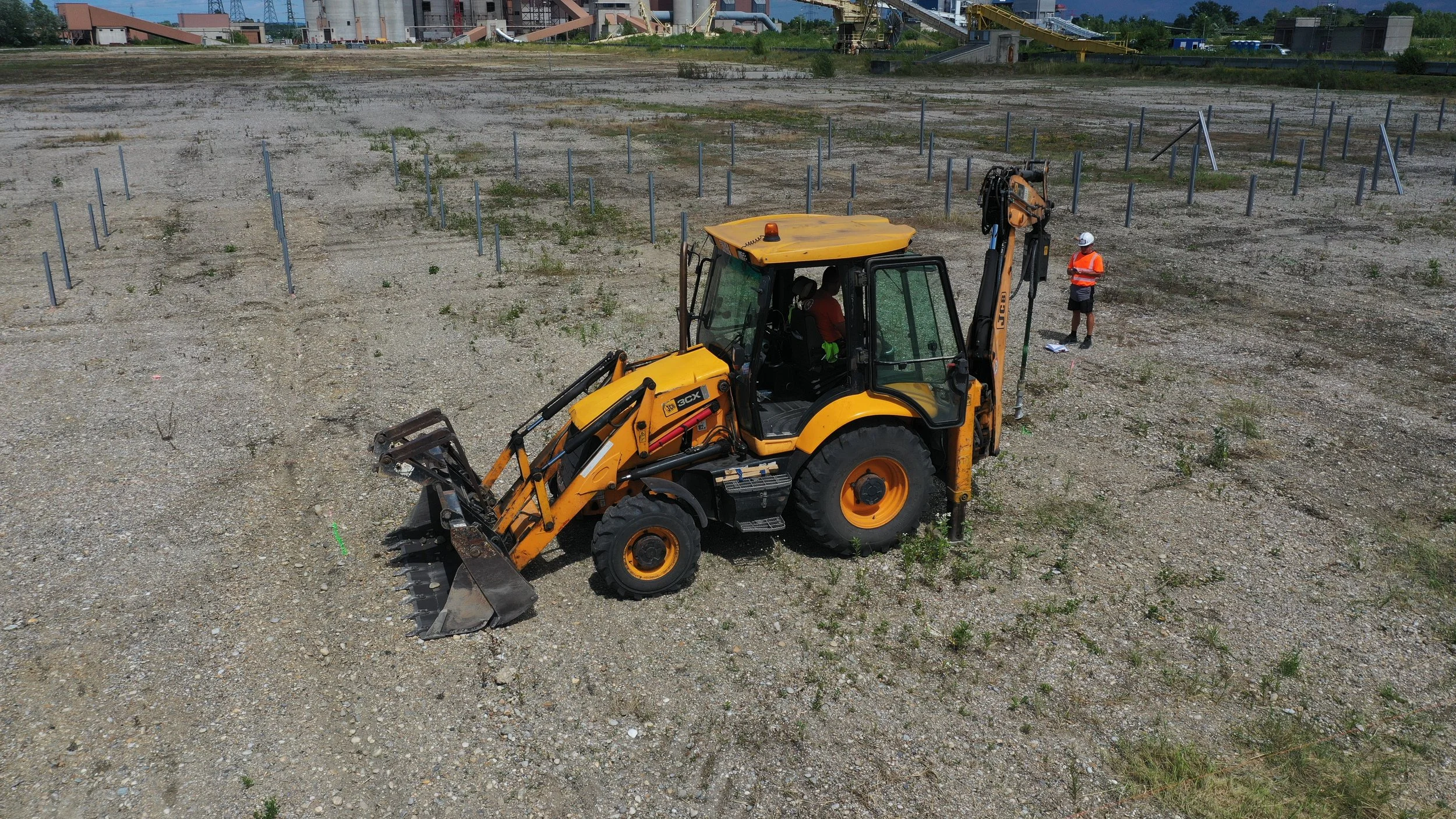 A yellow backhoe loader operated by a person in a red shirt is on a construction site with gravel ground. A worker wearing an orange safety vest and white helmet is standing nearby, holding a paper. In the background, there are industrial buildings and equipment.