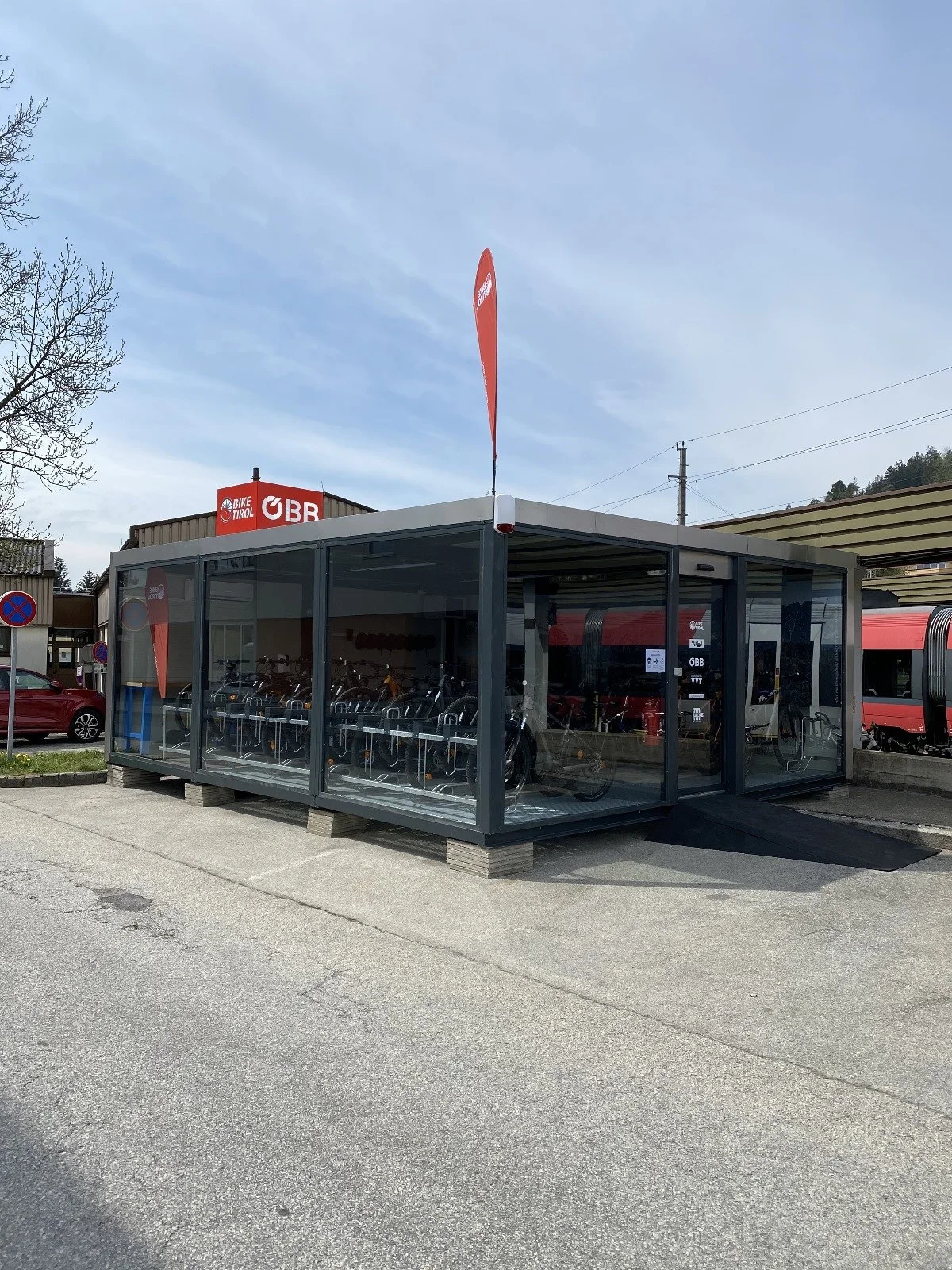 A glass-enclosed bike rental station with several bikes inside, situated outdoors near a street and buildings, with a flag and a sign for Bike Tirol and ÖBB visible.