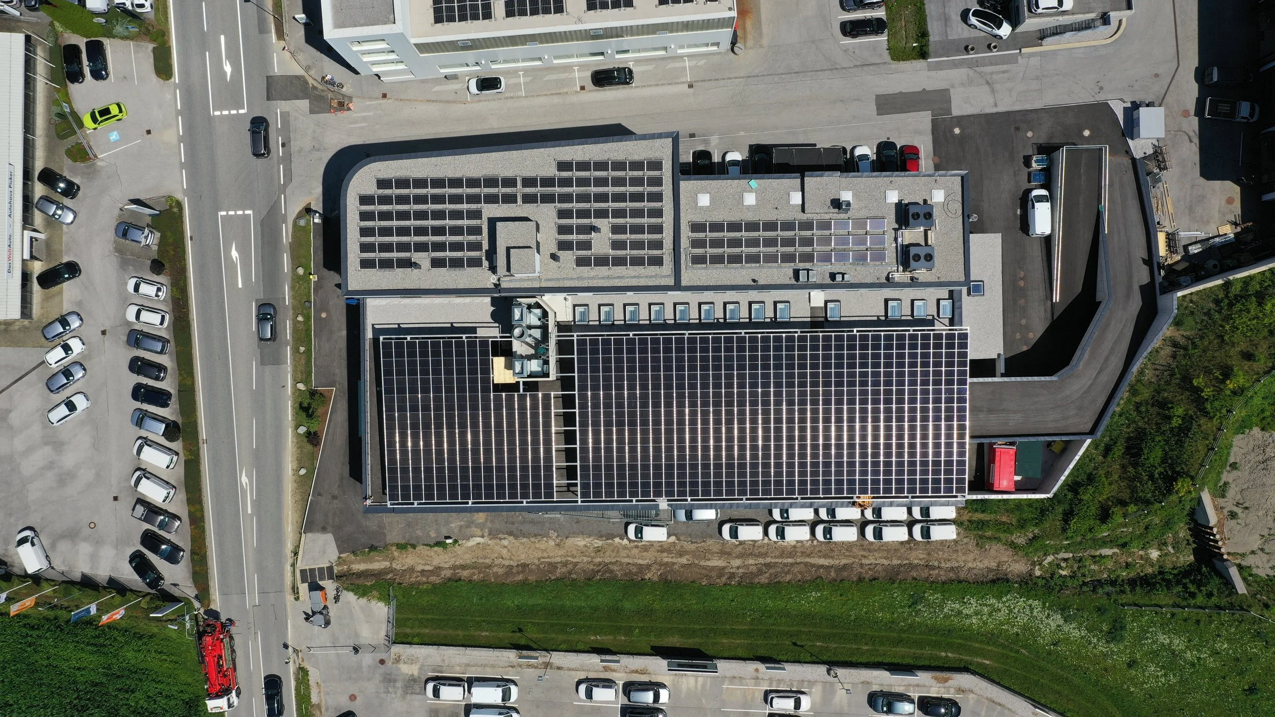 Légi felvétel egy épületről, amelynek tetején napelemek találhatók és egy carport, körülötte parkolókkal tele autókkal és szomszédos utakkal
