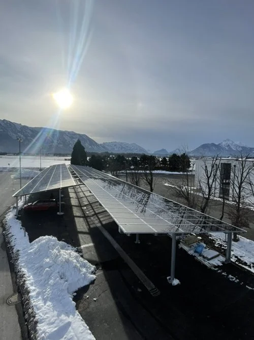 Solar panels on a rolling landscape with snow, mountains in the background, and the sun shining through a clear sky.