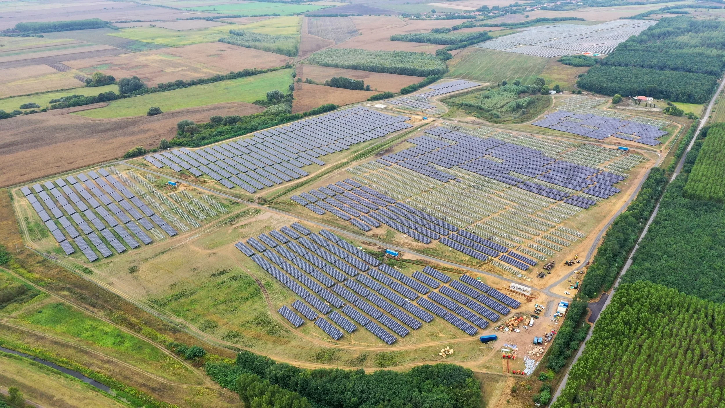 Aerial view of a large solar farm with numerous solar panels arranged in rows on a rural landscape, surrounded by farmland and greenery.