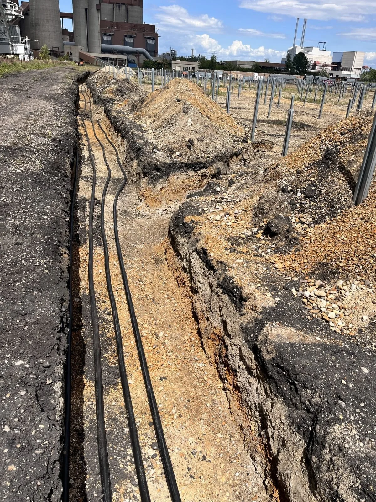 A construction site with a deep trench showing underground pipes, surrounded by piles of dirt and gravel, with an industrial building in the background.