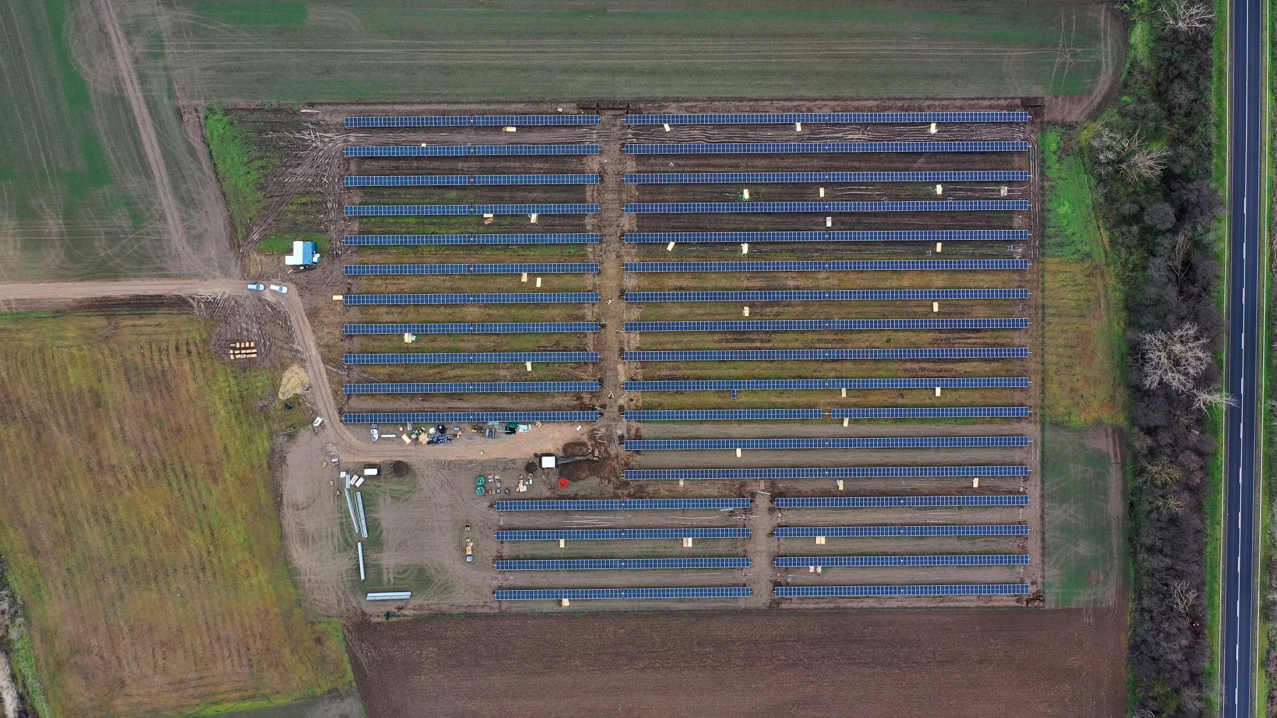 An aerial view of a large solar farm with rows of solar panels, dirt pathways, a few vehicles, and adjacent farmland and a highway on the right side.
