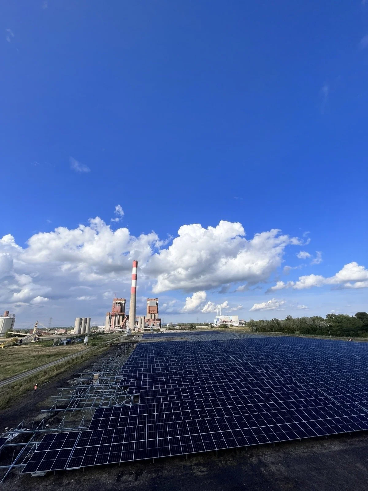 A solar power farm with solar panels in the foreground and an industrial facility with smokestacks in the background under a blue sky with scattered clouds.