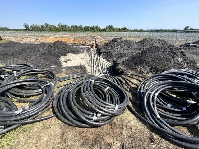 Electrical cables coiled on the ground at a construction site with underground pipes and piping trenches in the background.