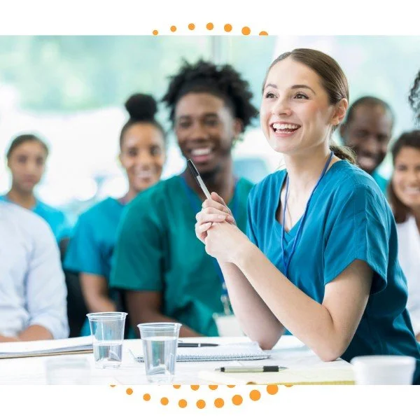 Group of healthcare professionals in scrubs smiling during a meeting or training session, with a female nurse in the foreground holding a pen.