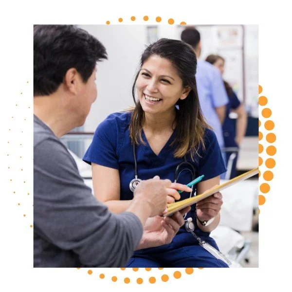 A female nurse wearing blue scrubs and a stethoscope engaging in a conversation with a male patient in a hospital or clinic setting.