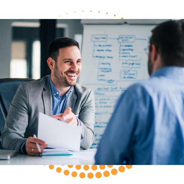 Two men in a business meeting, one smiling and holding papers, with a whiteboard and flowchart in the background.