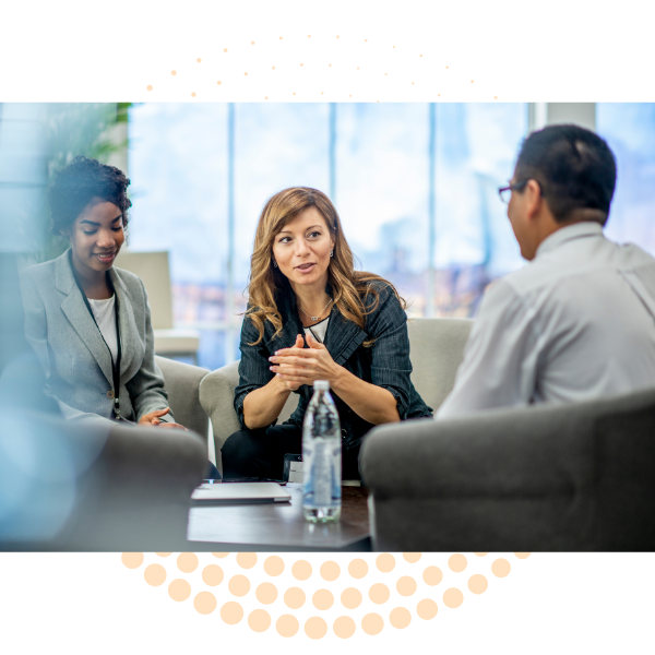 Three people sitting in a meeting room, engaged in conversation. One woman with long hair and a blazer is speaking, while a man with glasses and a white shirt listens, and another woman with short hair and a blazer looks on. There is a bottle of water on the table.