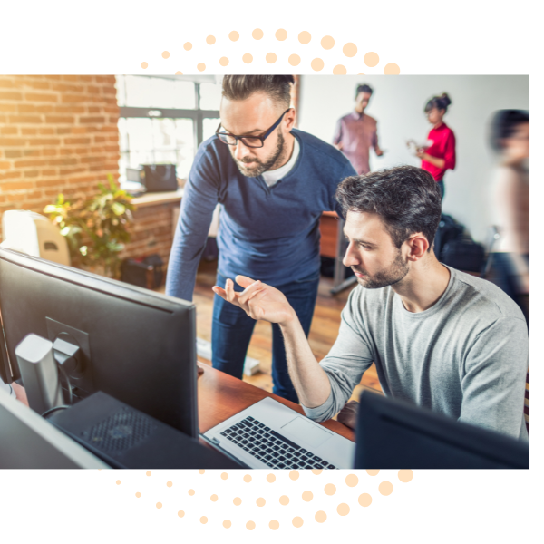 Two men working together at a computer in an office with others in the background.