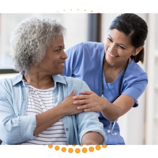 A healthcare worker or nurse smiling and touching the shoulder of an elderly woman who is smiling back, in a medical setting.