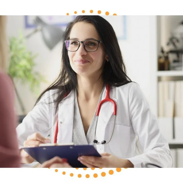 A female doctor with glasses and a stethoscope talking to a patient in a medical office.