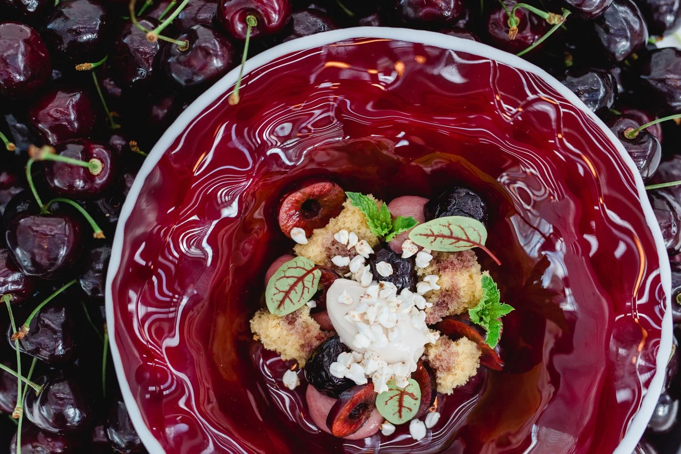 Ein Dessert mit roten Johannisbeeren, Früchten, Minzblättern, Nüssen sowie einer roten Gelee-Schicht in einer weißen Schale, umgeben von frischen Johannisbeeren.