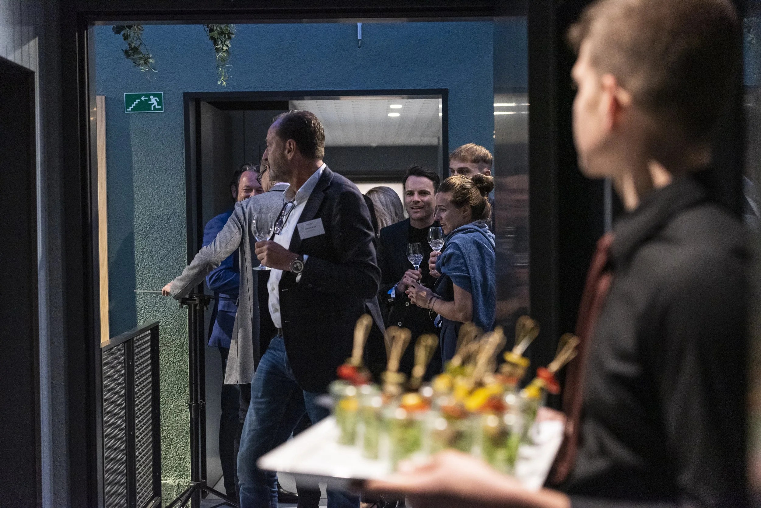 Menschen bei einer Dinner-Party, einige mit Weingläsern, im Hintergrund eine Person, die mit einem Tablett voller Häppchen steht.