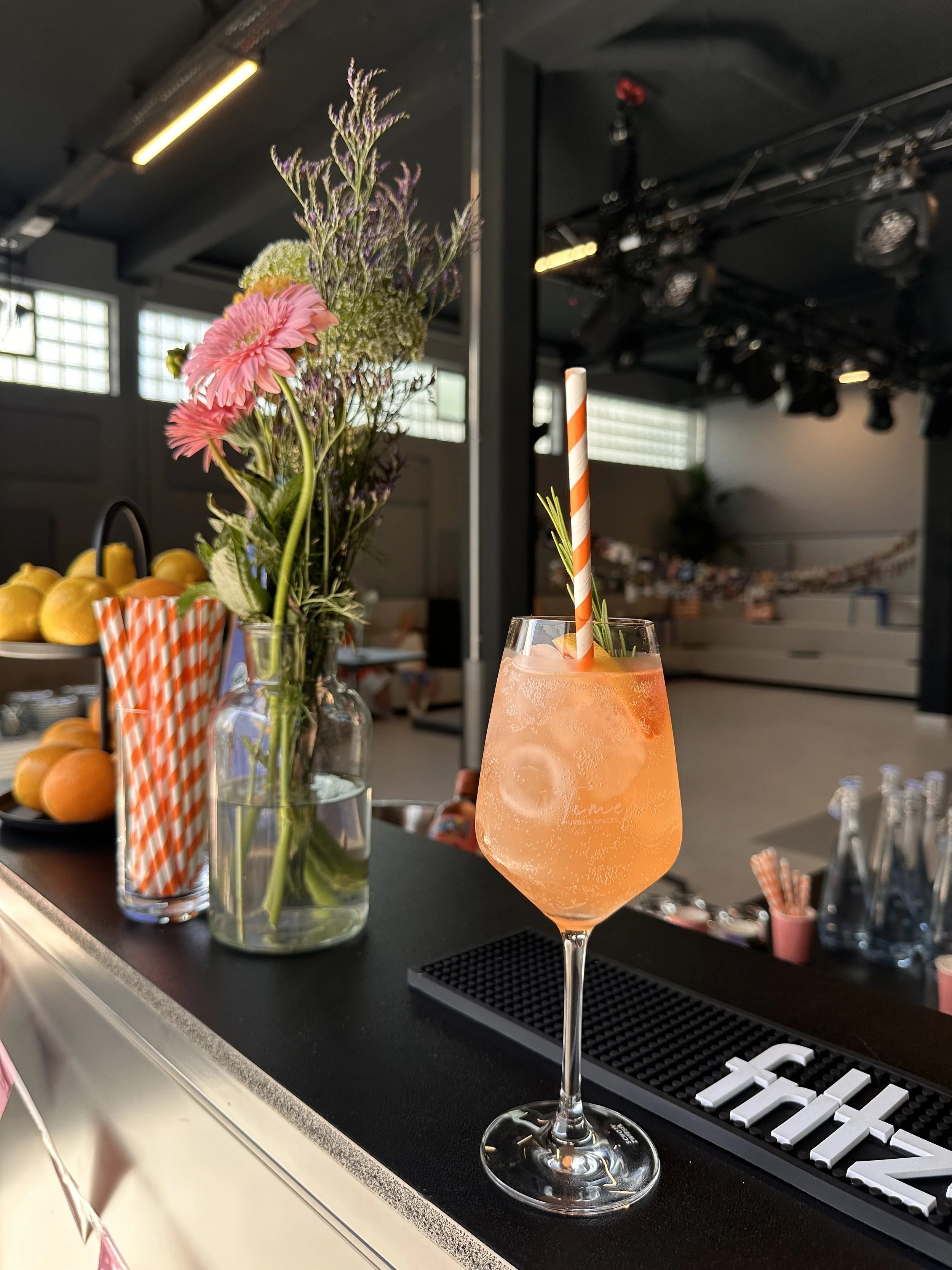 Ein Orangefarbener Cocktail mit Eis in einem Weinglas, mit einem gestreiften Trinkhalm und einer kleinen Kräuterdekoration. Im Hintergrund eine Vase mit Blumen und eine Schale mit Zitrusfrüchten in der Factory41 der Urban Spaces.