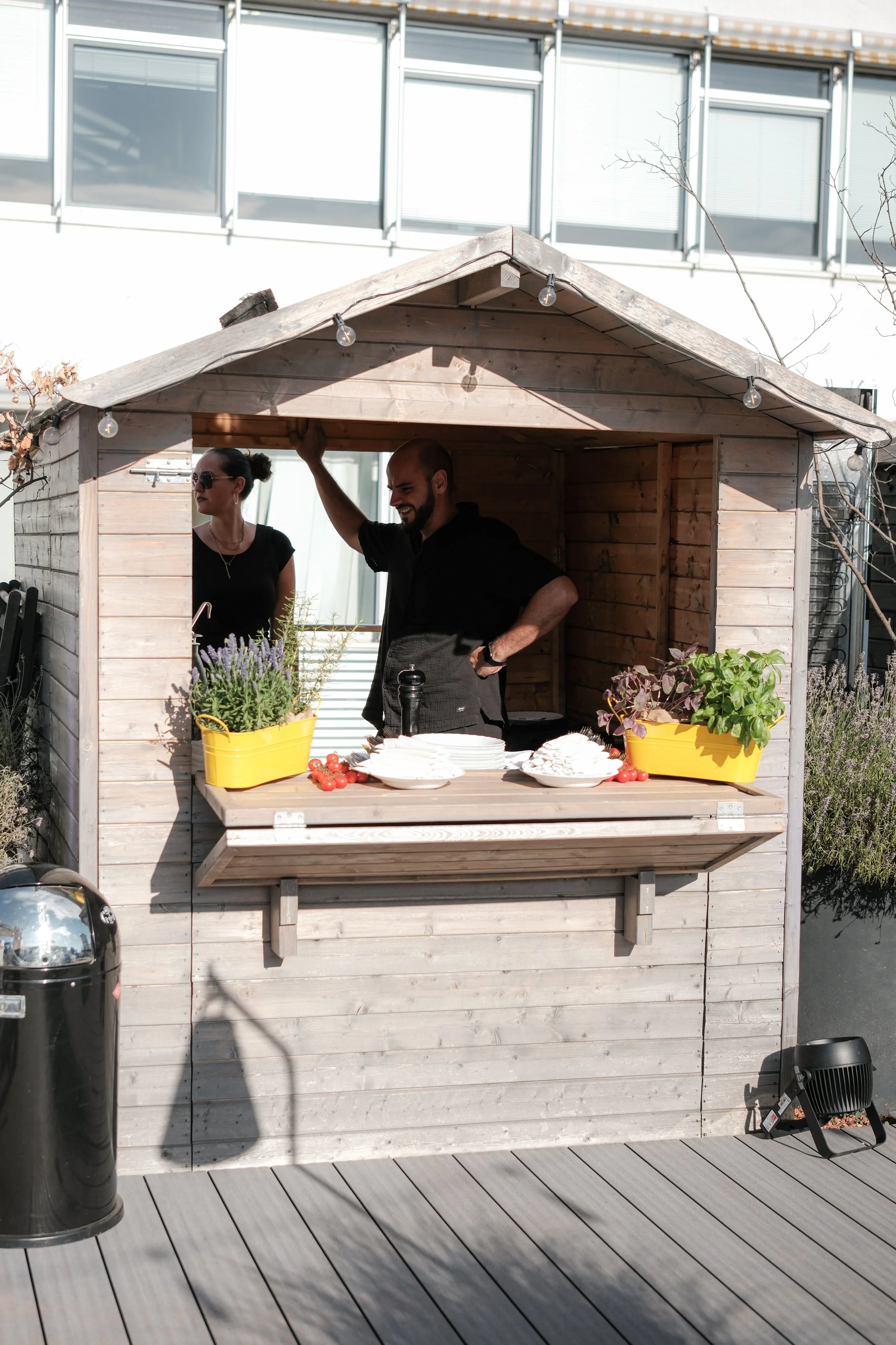 Ein Mann und eine Frau stehen in einem kleinen Holzpavillon im Freien der Urban Spaces, umgeben von Pflanzen, mit einem Tisch voller Teller, Tomaten und Kräuter, bei Tageslicht.
