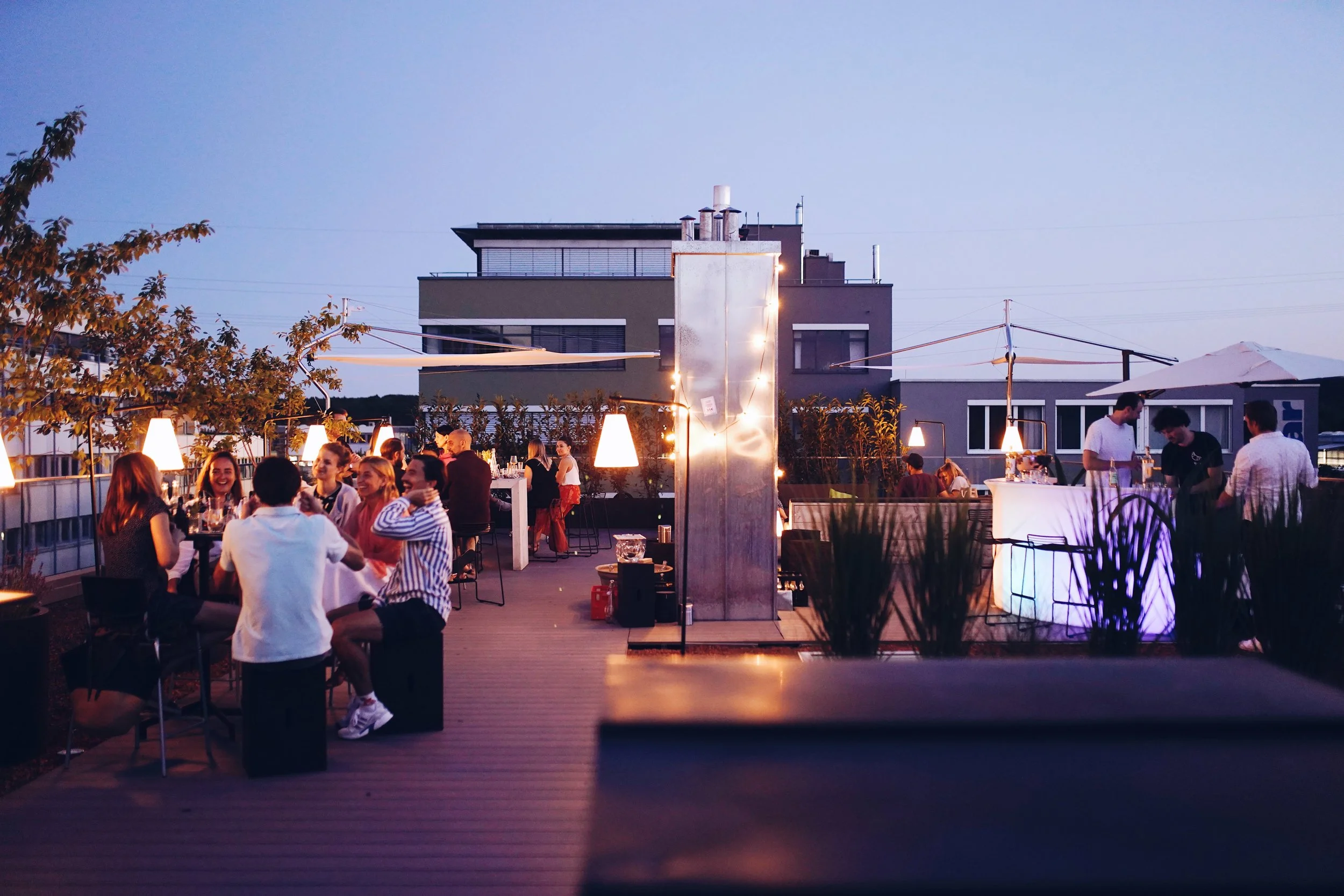 Menschen genießen einen Abend auf der Dachterrasse  der Urban Spaces mit moderner Beleuchtung und Blick auf den Himmel, draußen bei Dämmerung.