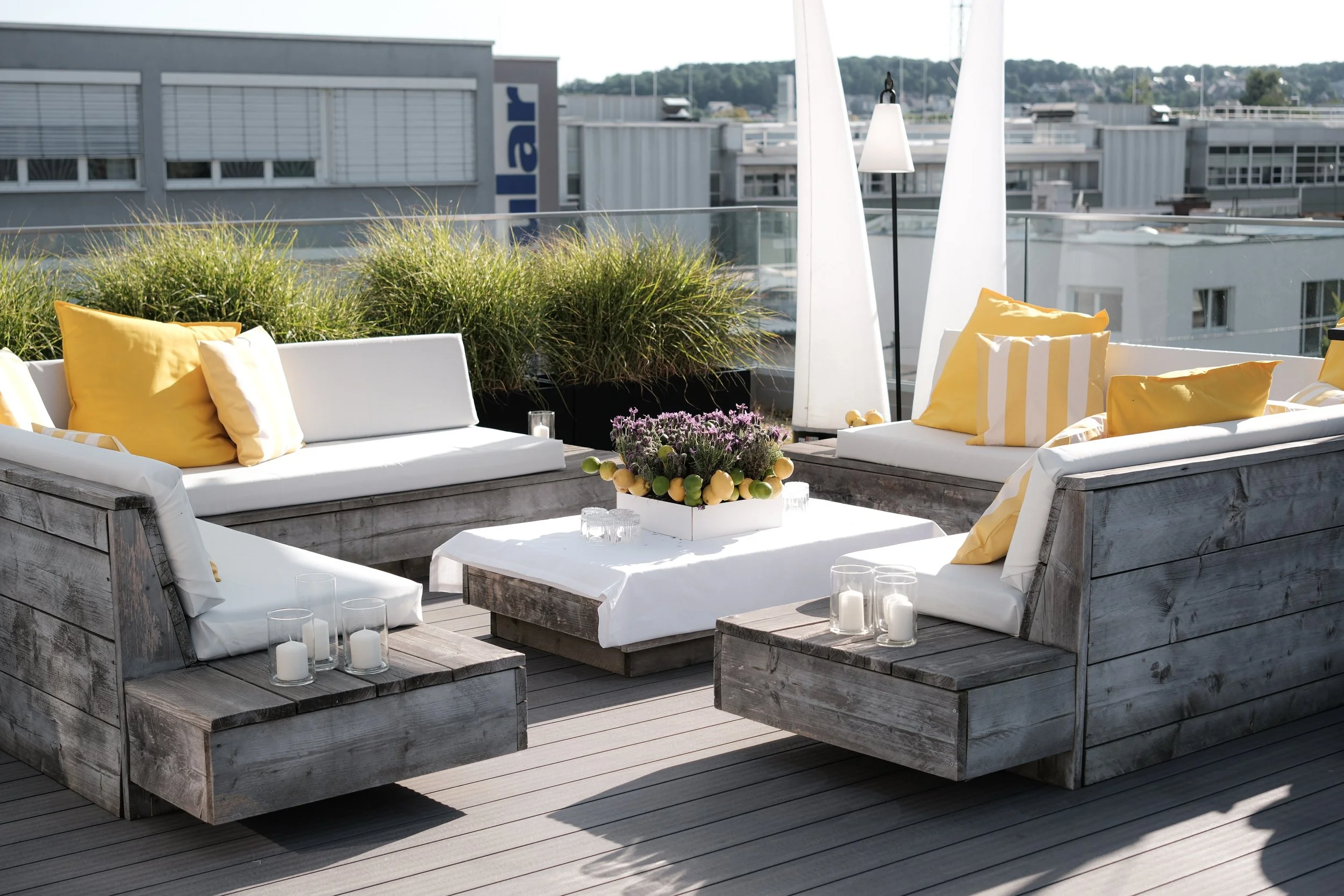 Geräumige Dachterrasse in den Urban Spaces mit Sitzbereich, bestehend aus Holzmöbeln, weißen Polstern, gelben Kissen, einem weißen Tisch mit Blumen, Kerzen und einer stylischen Stehlampe, mit Blick auf Stadtgebäude und Himmel.