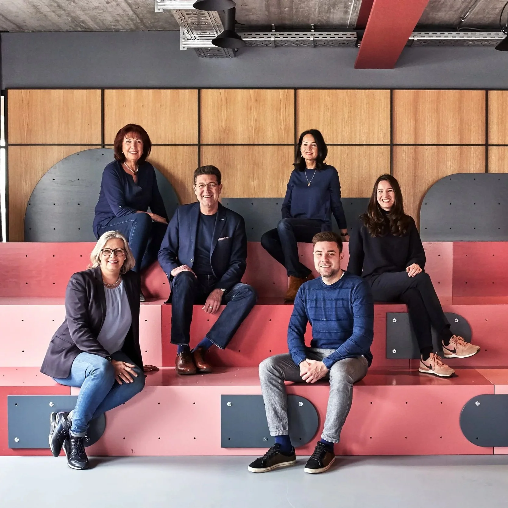 Das Team der Urban Spaces sitzt auf einer modernen Sitztribüne im Worklba der Urban Spaces vor einer Holz-Wand in einem Raum mit industriellem Deckenstil.
