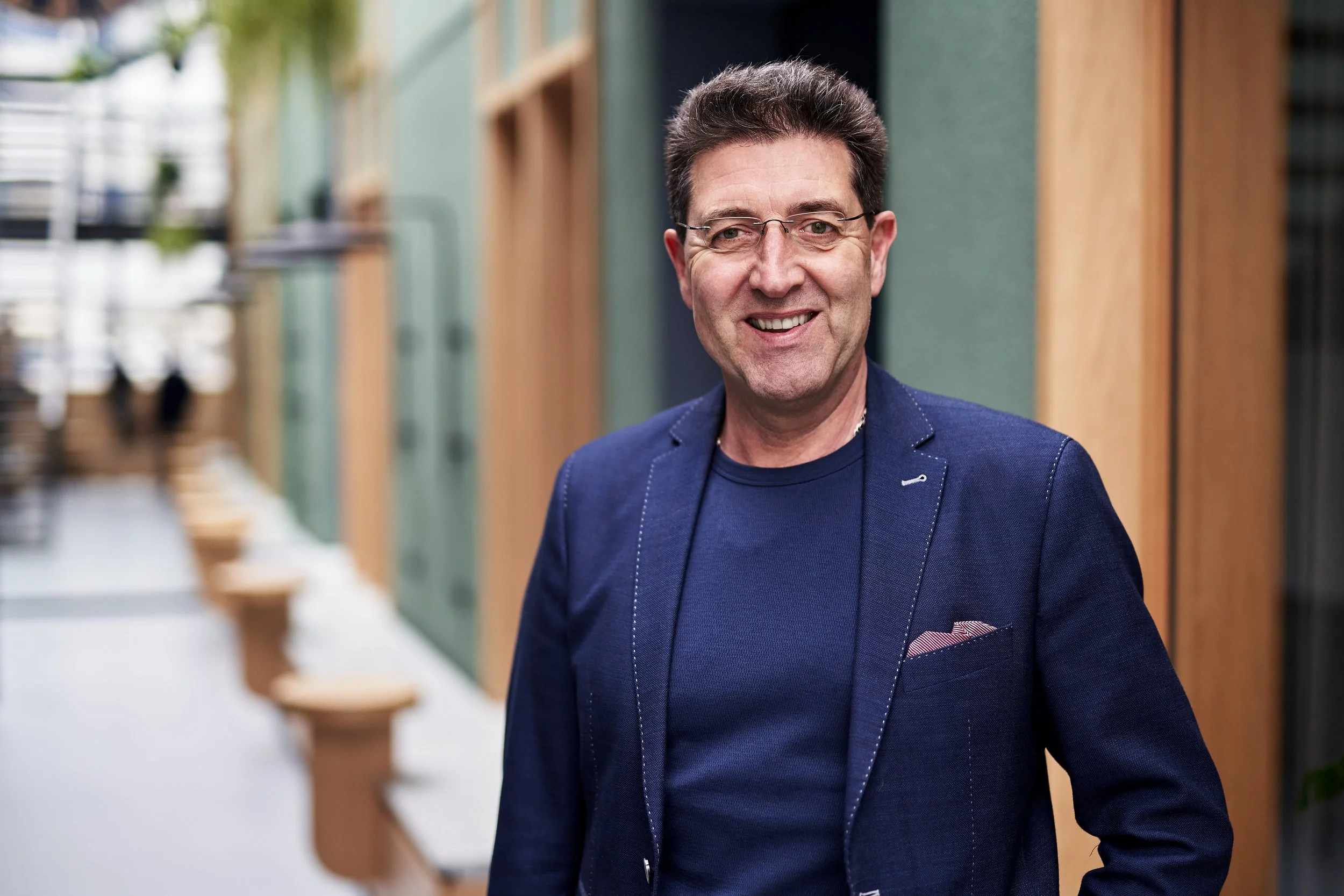 Ralf Schuck in einer blauen Jacke und Brille steht in einer modernen, hellen Indoor-Umgebung mit Holzmöbeln und grünen Wänden.