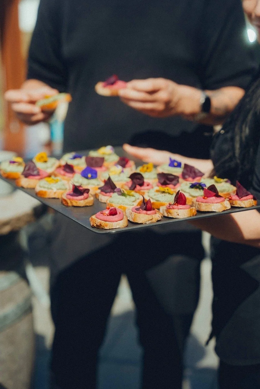 Eine Person hält eine Platte mit kleinen, bunten Canapés, die mit verschiedenen Blüten und Cremes garniert sind.
