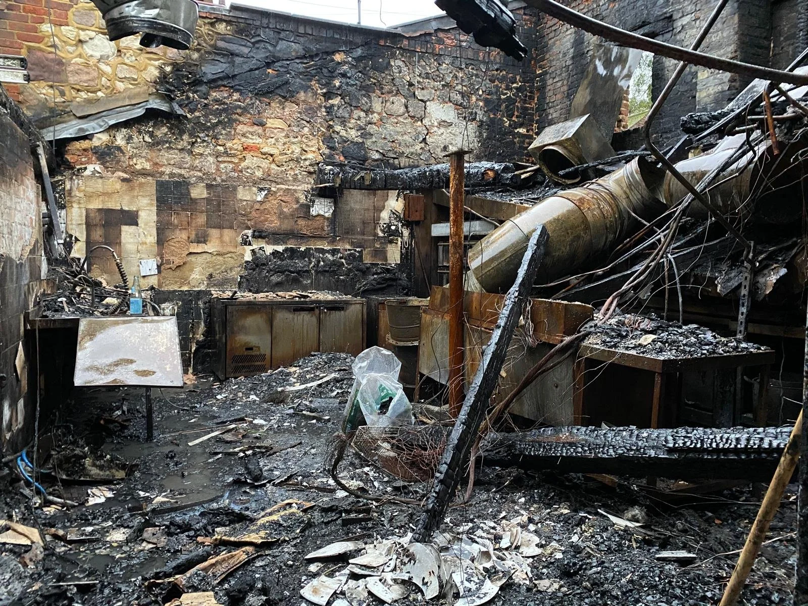 A room that has been severely damaged by fire, with burnt walls, charred debris on the floor, and damaged equipment and appliances.