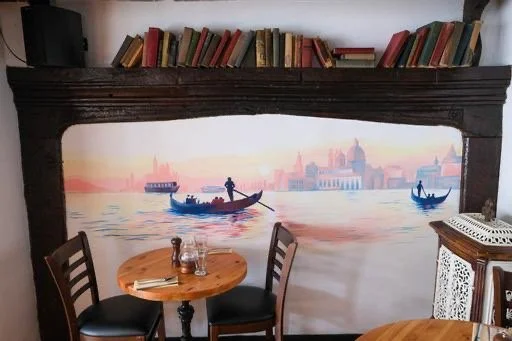 Wall mural of a river scene with boats, books on a shelf above, two wooden tables and chairs in the foreground, and a white lace chair on the right.