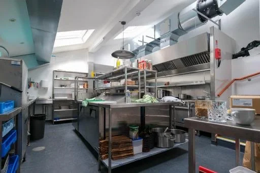 Commercial kitchen with stainless steel counters, shelves, and cooking equipment, including a ventilation hood, mixer, and various utensils and containers.