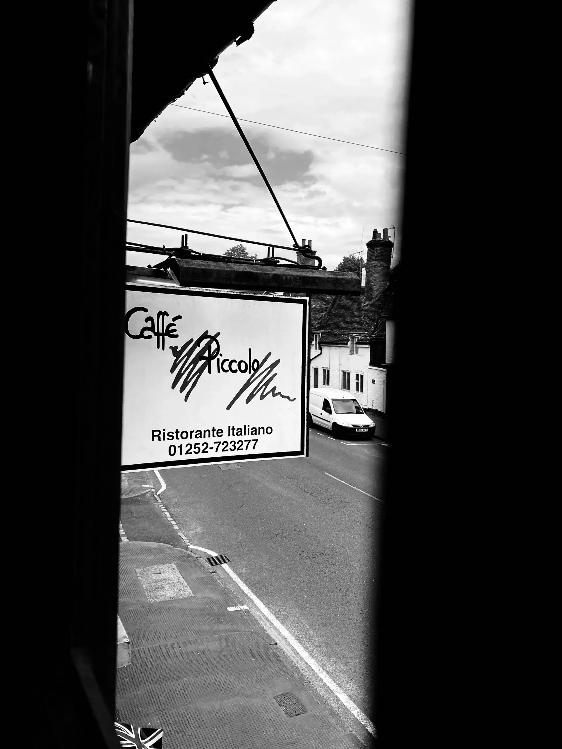 View of a street scene through a window with a restaurant sign for 'Caffé Piccolo' and its phone number, with a white van parked on the street and buildings with chimneys in the background, in black and white.