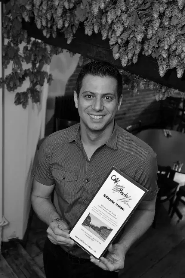 A smiling man holding a certificate in a restaurant setting with hanging dried chili peppers overhead.