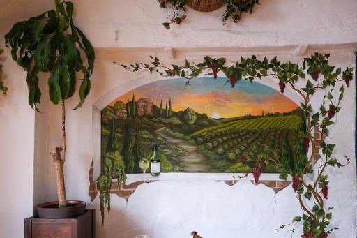 Mural of a vineyard landscape painted on a wall, with a sunset sky, rolling hills, and grapevines, surrounded by hanging plant leaves.