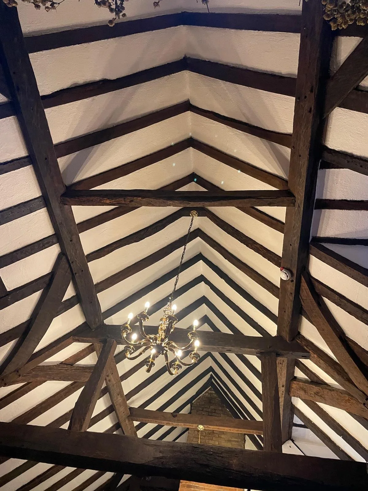 Interior view of a wooden ceiling with exposed dark wood beams and white plaster. A chandelier hanging from the ceiling lights up the room.